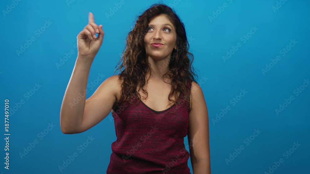 Fototapeta premium Young hispanic woman pointing index finger upward with raised arm in studio; confidence curiosity discovery.