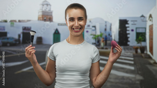 Fotografi Young woman with short buzz cut holding a makeup brush in left hand and a beauty sponge in right on a street crosswalk, smiling and posing; playful confidence