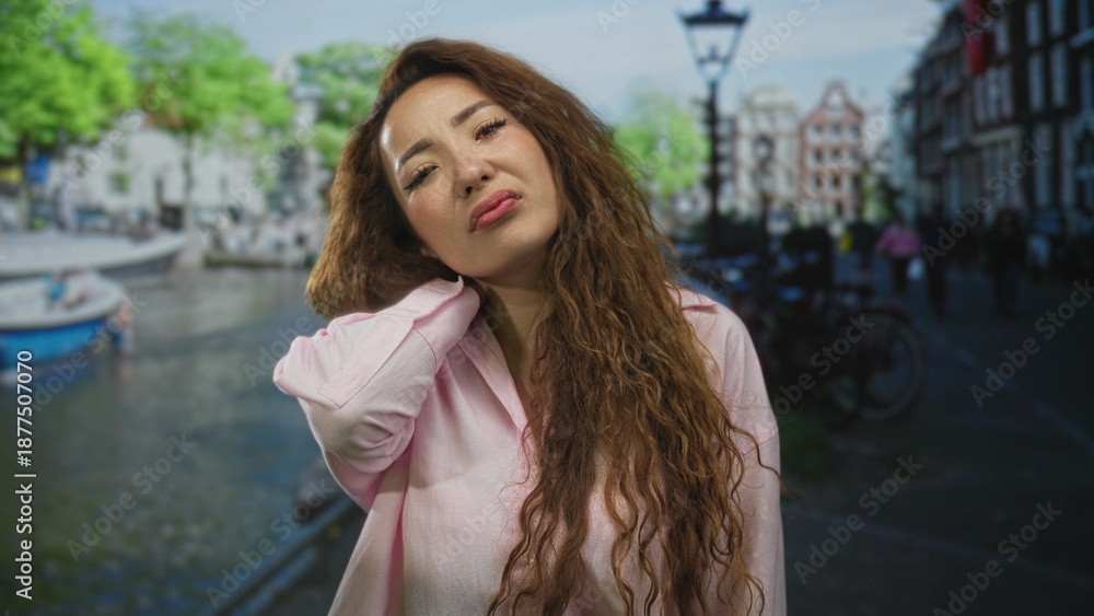 Fototapeta premium Woman with hand on neck on amsterdam street by canal boats and bikes, showing neck pain; discomfort.