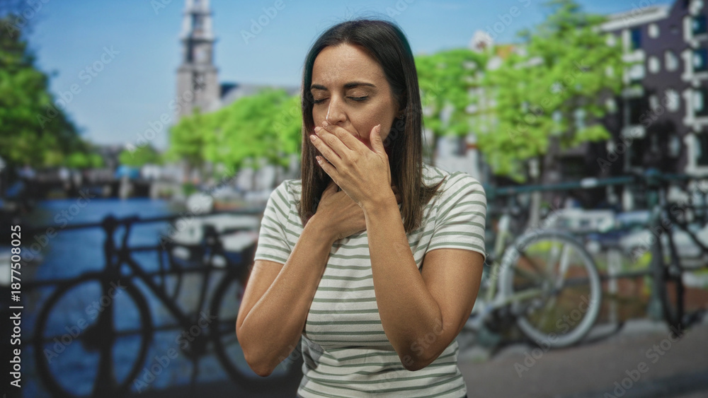 Fototapeta premium Woman young hispanic holding throat with both hands beside a parked bicycle on a street by a canal and bridge; breathing difficulty anxiety.