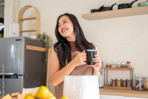 Smiling asian woman holding coffee mug standing at kitchen cooking counter with fruit and toaster.