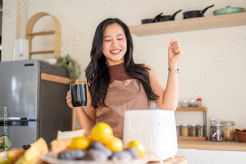 Pretty asian woman holding coffee mug dancing aside kitchen cooking counter with fruits and toaster.