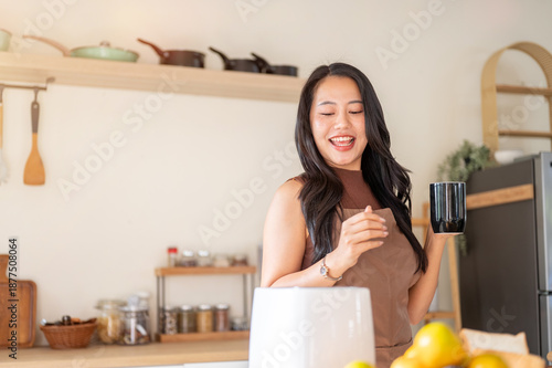 Happy asian woman holding coffee mug dancing aside kitchen cooking counter with fruits and toaster.