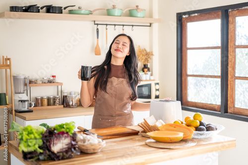 Happy asian woman holding coffee mug standing at kitchen cooking counter with vegetables and fruit.