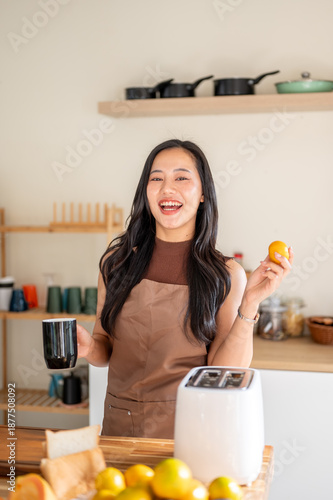 Pretty asian woman holding coffee mug and orange standing aside toaster on kitchen cooking counter.