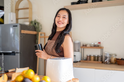Pretty asian woman holding a coffee mug while standing aside toaster and fruits on kitchen counter.