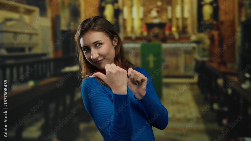 Fototapeta premium Young woman in blue sweater points thumbs behind while smiling inside an atmospheric church, capturing a playful and engaging gesture in a religious setting.