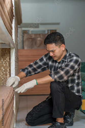 Asian adult male woodworking business owner kneeling near wood panels and reviewing material condition, professional carpenter performing inventory inspection inside interior furniture material store
