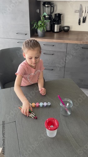 Little girl putting colorful paints and brushes into a glass jar. Organizing Easter art supplies for creative session.