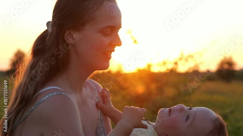 A woman gently holds her baby close as the sun sets in the background. They stand in an open field surrounded by tall grass and trees