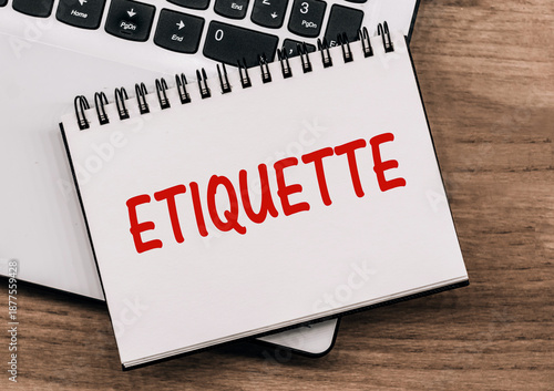 A white notebook with a red Etiquette written on it sits on a wooden desk near a closed laptop.