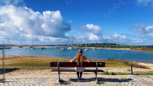 woman sitting on bench looking at sea and fishing boat, Portugal, Algarve