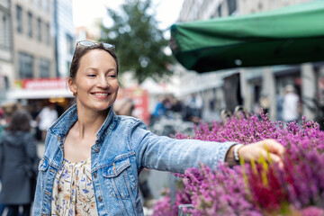 Young adult joyful woman choose to buy colorful potted flowers at outdoor market stall. Female person at sale of blooming plants in pots at botanical street German store on sunny spring autumn day