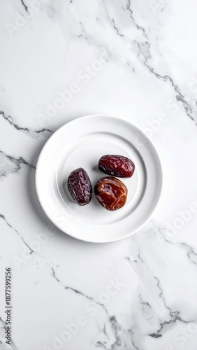 Three glistening dark red dates arranged on a small white plate on a white marble surface with subtle gray veining creating a minimalist food still life.