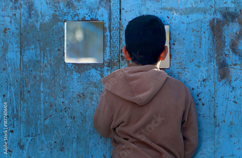 A child peers through a crack in a school door in a working-class neighborhood, watching children playing football in the evening after school hours.
