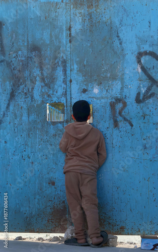 A child peers through a crack in a school door in a working-class neighborhood, watching children playing football in the evening after school hours.