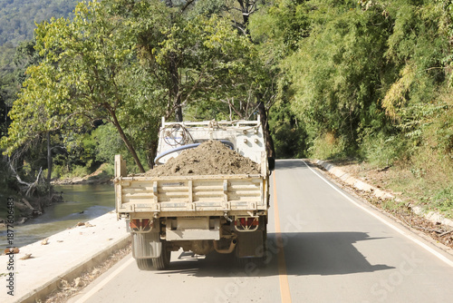 White pickup truck driving on rural road transport heavy load of soil along scenic route with green nature and peaceful river flow on sunny day