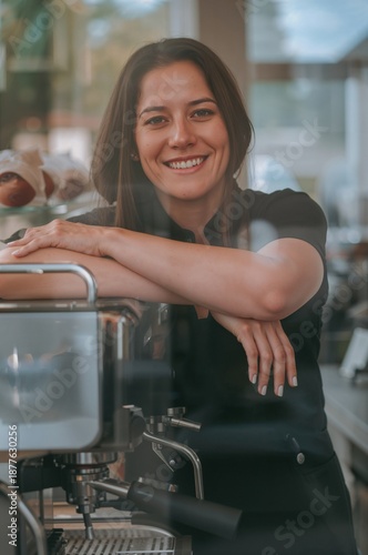 Friendly female barista smiles at the camera while leaning on a professional espresso machine inside a modern cafe with soft natural light and blurred background details