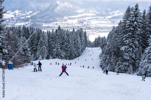 Numerous skiers descending a snowy piste towards the valley, surrounded by dense pine forests.