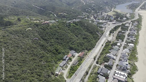 Drone flies backwards looking down at paragliders over coastal highway on a sunny day on the Garden Route in Wilderness, South Africa
