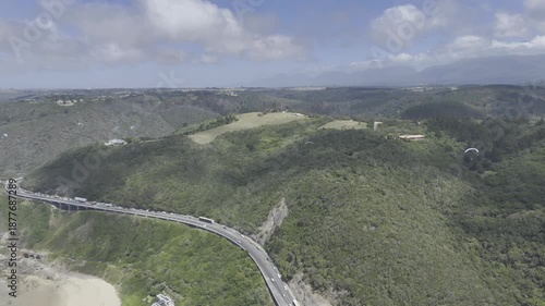 Drone flies east high over coastal highway with paragliders above on a sunny day on the Garden Route in Wilderness, South Africa