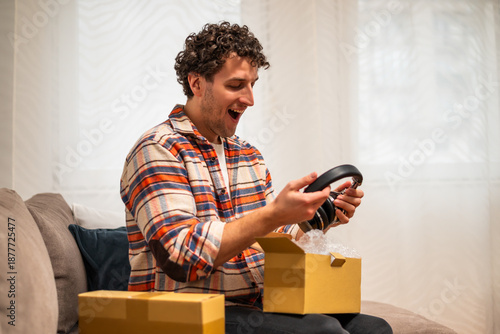 Wallpaper Mural Happy young man smiling and opening delivered package with headphones while sitting on sofa at his home. Image captures joy and convenience of home shopping and receiving order.	 Torontodigital.ca
