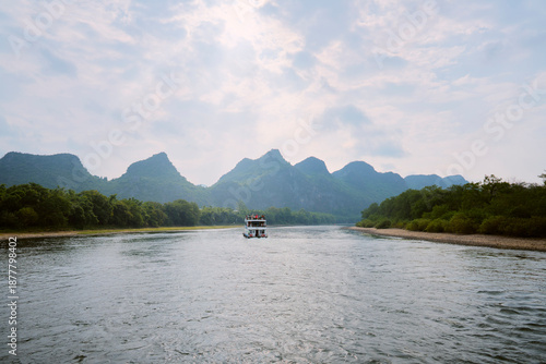 A tranquil riverboat cruises along a wide river with lush green hills in the background. Li River, Li Jiang, Guangxi, China