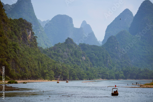 Serene river flows through lush mountains under a clear blue sky with boats on water.  Li River, Li Jiang, Guangxi, China