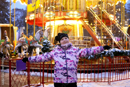 Happy girl in pink jacket posing near illuminated carousel, child enjoying winter evening at bright amusement park fairground during holidays