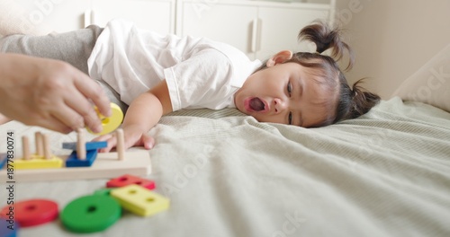 Young Asian toddler with pigtails lies mid yawn on a light bed as an adult hand arranges wooden shape sorter pieces in a bright modern living room in daylight.