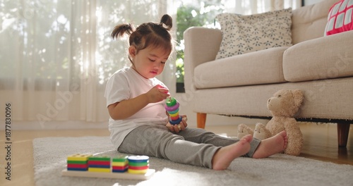 A two year old Asian girl sits barefoot on a soft gray rug, stacking ring toys near sheer curtains, a beige sofa, patterned pillows, and a plush teddy bear.