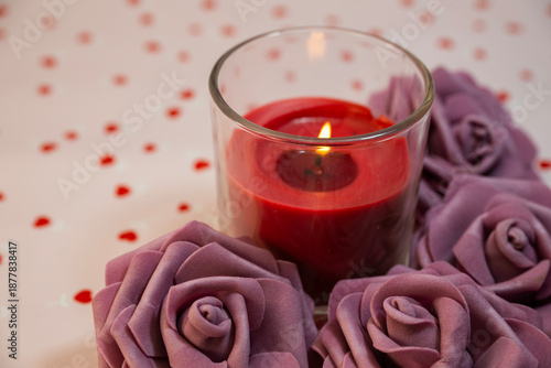 burning red candle in a glass jar, surrounded by large, soft mauve roses on a surface scattered with red heart confetti.