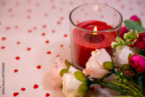 A romantic close-up captures a lit red candle in a glass holder nestled next to pink roses on a pale pink surface scattered with red heart confetti.
