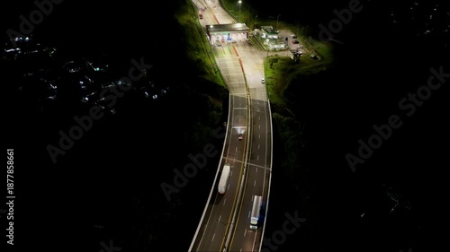 Illuminated toll road and winding highway cutting through dark landscape at night with vehicles passing through, viewed from a high angle.