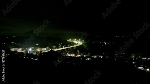 Curving road illuminated by street lights cutting through dark hillside landscape at night, captured from a drone perspective.