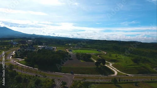 Green countryside landscape with winding road and open fields stretching toward distant hills, seen from aerial view.