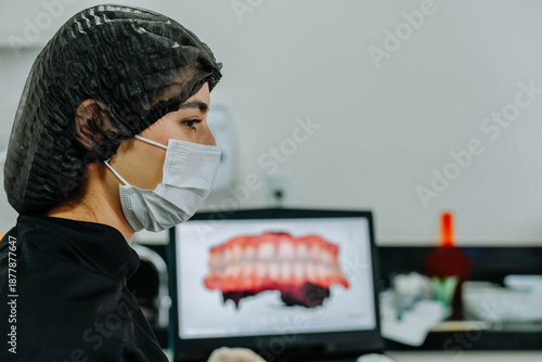 Female dentist wearing protective equipment while working in a dental clinic with dental images visible on a computer screen.