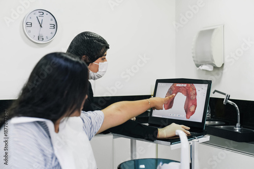 Dentist reviewing dental images on a computer while a female patient sits nearby during a consultation in a dental clinic.