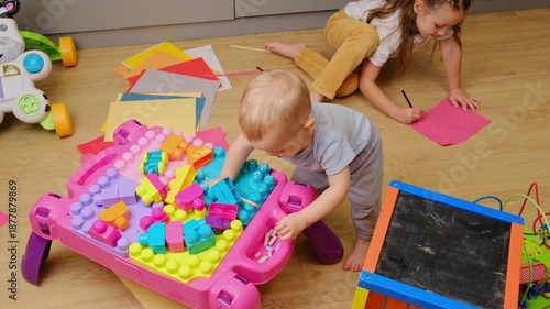 Adorable little brother having fun at home playing together on floor with wooden toys, little girl drawing coloring pictures in paper album 