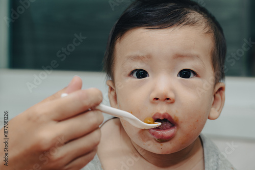 Wallpaper Mural An expressive close-up of a content Asian baby enjoying mealtime, being fed by a spoon. A curious baby eating with a spoon. A joyful Asian infant being spoon-fed, showing a messy face and big eyes. Torontodigital.ca
