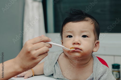 Wallpaper Mural An expressive close-up of a content Asian baby enjoying mealtime, being fed by a spoon. A curious baby eating with a spoon. A joyful Asian infant being spoon-fed, showing a messy face and big eyes. Torontodigital.ca