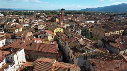 Aerial 4K view of Lucca skyline, flying from the historic Clock Tower (Torre delle Ore) towards Guinigi Tower with oak trees, Tuscany, Italy.