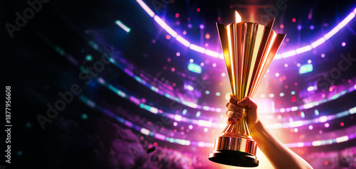 Wide banner of a hand holding a gold trophy cup against a blurred stadium background with vibrant neon lights, featuring copy space for victory celebration and championship concepts