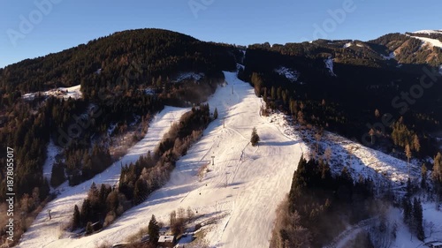 Aerial snowy alpine ski slope at sunset, golden light casting long shadows across groomed runs, dense evergreen forest flanking