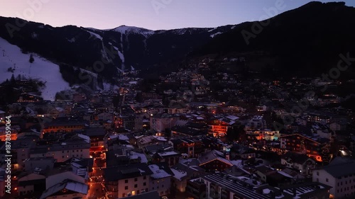 twilight alpine town under snow slopes with glowing streetlights and lit rooftops, ski runs visible as pale ribbons, aerial drone