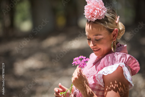 Portrait of young woman, beautiful, blonde, with typical Andalusian flamenco costume to go on pilgrimage with a small shawl and pink flower, looking at the violet flowers in her hands.