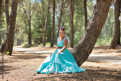 Young, beautiful, blonde woman wearing a typical Andalusian flamenco dress in light blue for going to a pilgrimage, holding a stick in her hand, sitting and resting on the trunk of a tree.