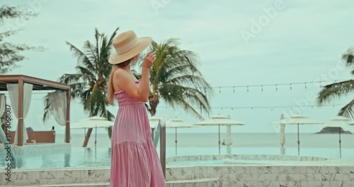 Woman in straw hat and pink dress standing by a luxurious infinity pool, overlooking the serene ocean and tropical beach with palm trees. Luxury vacation, tropical travel, relaxation, summer lifestyle
