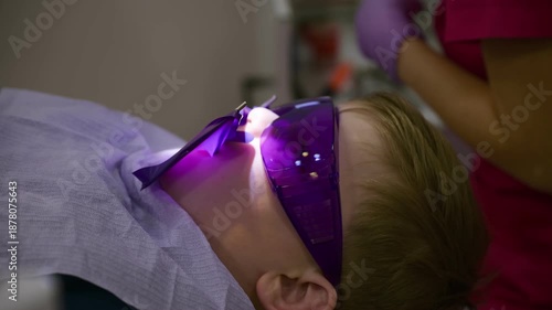 A young child rests in a dental chair, wearing protective glasses while a dentist performs an important slow motion oral treatment in a clinical setting.
