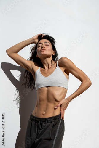 Athletic adult woman with defined abdominal muscles posing in sunlight against white wall, minimalist fitness lifestyle portrait, natural body shape, strength, confidence, and calm wellness concept